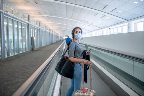 راكبة شابة تستخدم السلم الكهربائي في المطار.  لديها محفظتها وحقيبة أوراقها معها.  أنت ترتدي قناعًا للوجه لمنع انتشار الجراثيم.